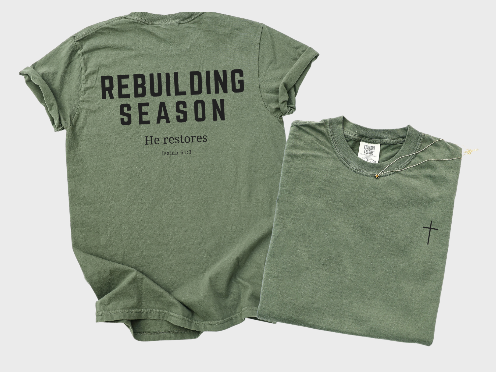 Plain white background with a moss colored t-shirt showing the back design that reads "Rebuilding Season He Restores Isaiah 61:3" and a cross on the front left chest area.