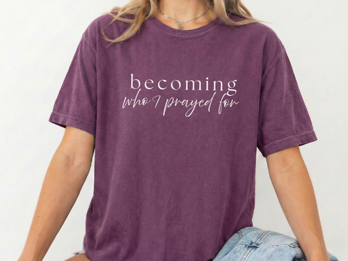 person sitting in front of a plain white background with a berry colored shirt with the text "becoming who i prayed for"